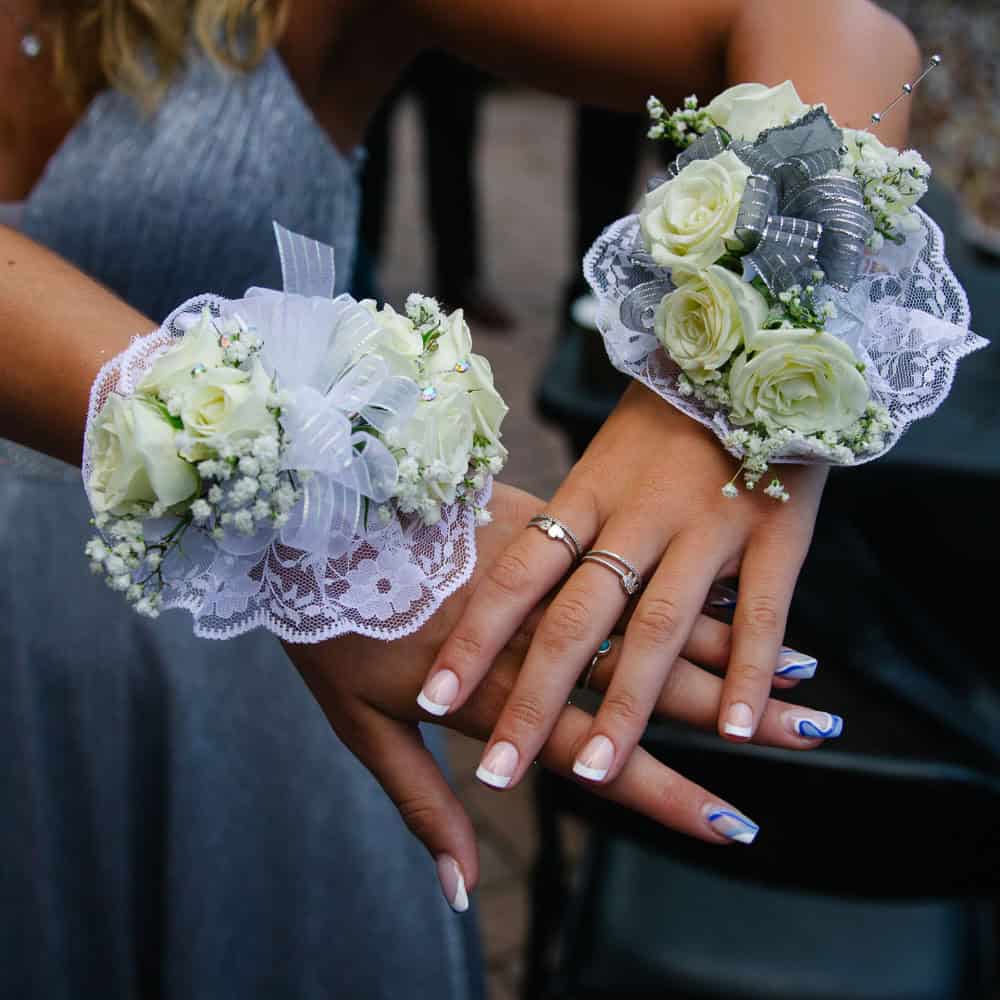 Two hands with white floral corsages, lace accents, and manicured nails resting atop each other, with a woman in a silver dress in the background.