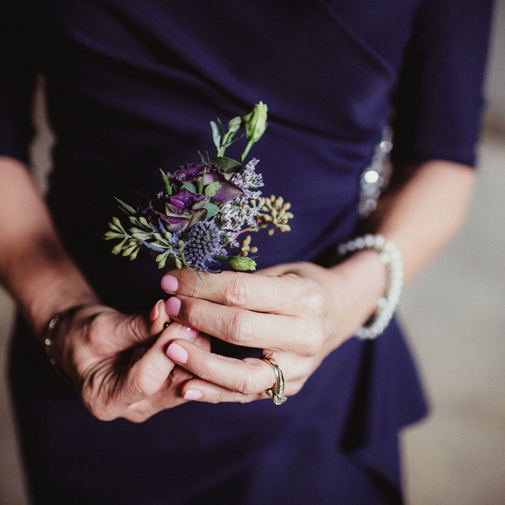 A woman in a dark blue outfit holds a small bouquet of purple and green flowers; they wear a ring and a pearl bracelet.