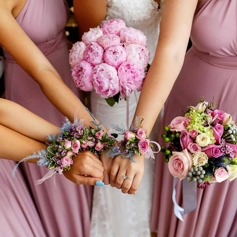 Three women in mauve dresses and a bride hold pink floral arrangements and wrist corsages, with their hands extended together in a celebratory gesture.