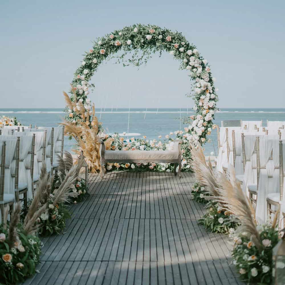 A floral wedding arch and bench are set up on a wooden aisle by the sea, with rows of white chairs and decorative greenery on each side.