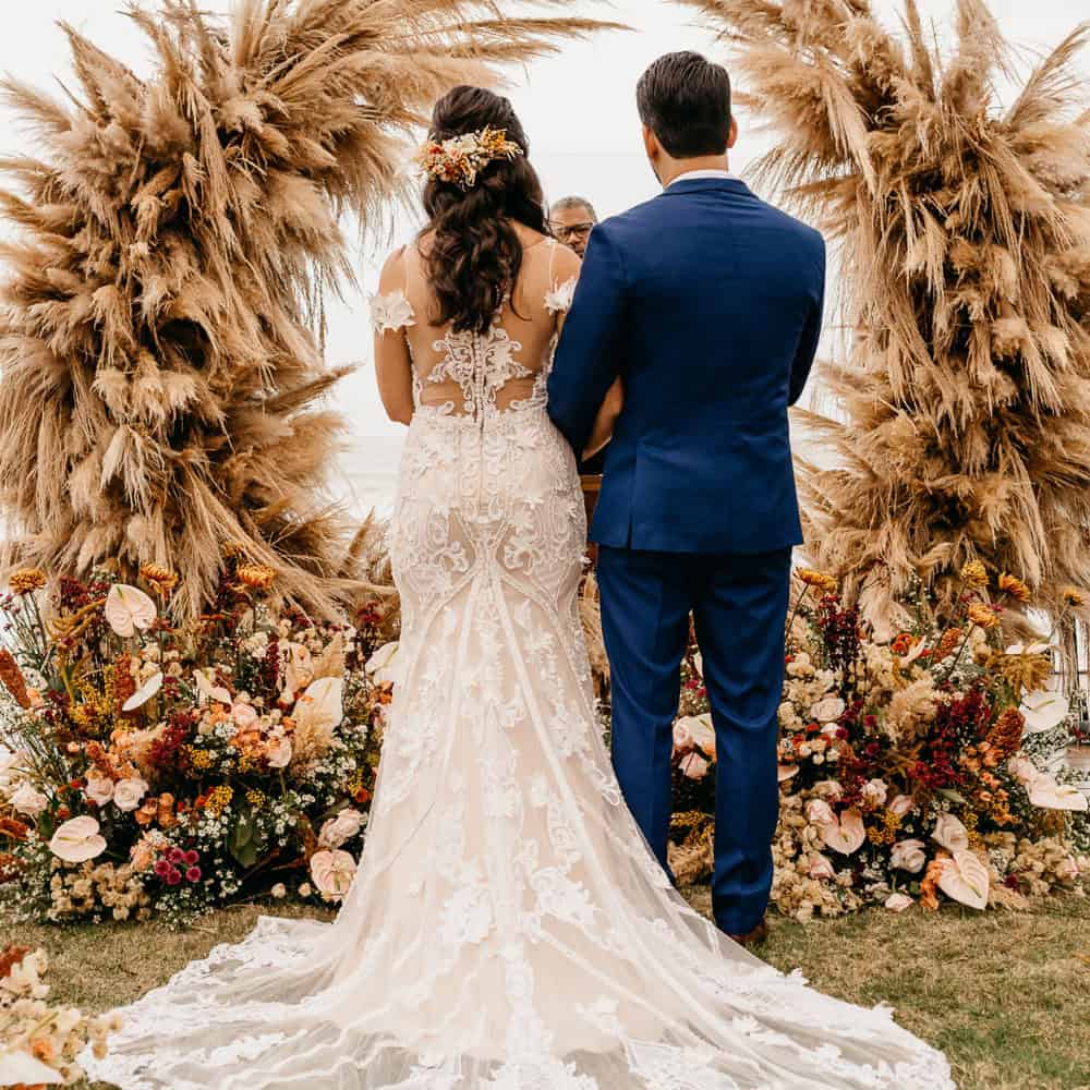 A bride and groom stand in front of a floral and pampas grass arch during an outdoor wedding ceremony, facing the officiant.