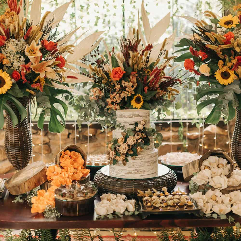 A decorated table displays a tiered white cake with floral accents, surrounded by vibrant flower arrangements and assorted sweets.