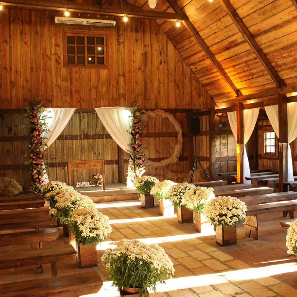 A rustic wooden barn decorated with white flowers and floral arches, set up with benches for a wedding ceremony. Sunlight streams in through the windows.