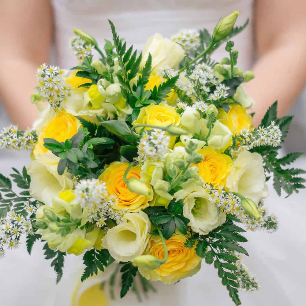 A close-up of a bouquet featuring yellow and white roses, small white flowers, green leaves, and ferns, held by a woman in a white outfit.