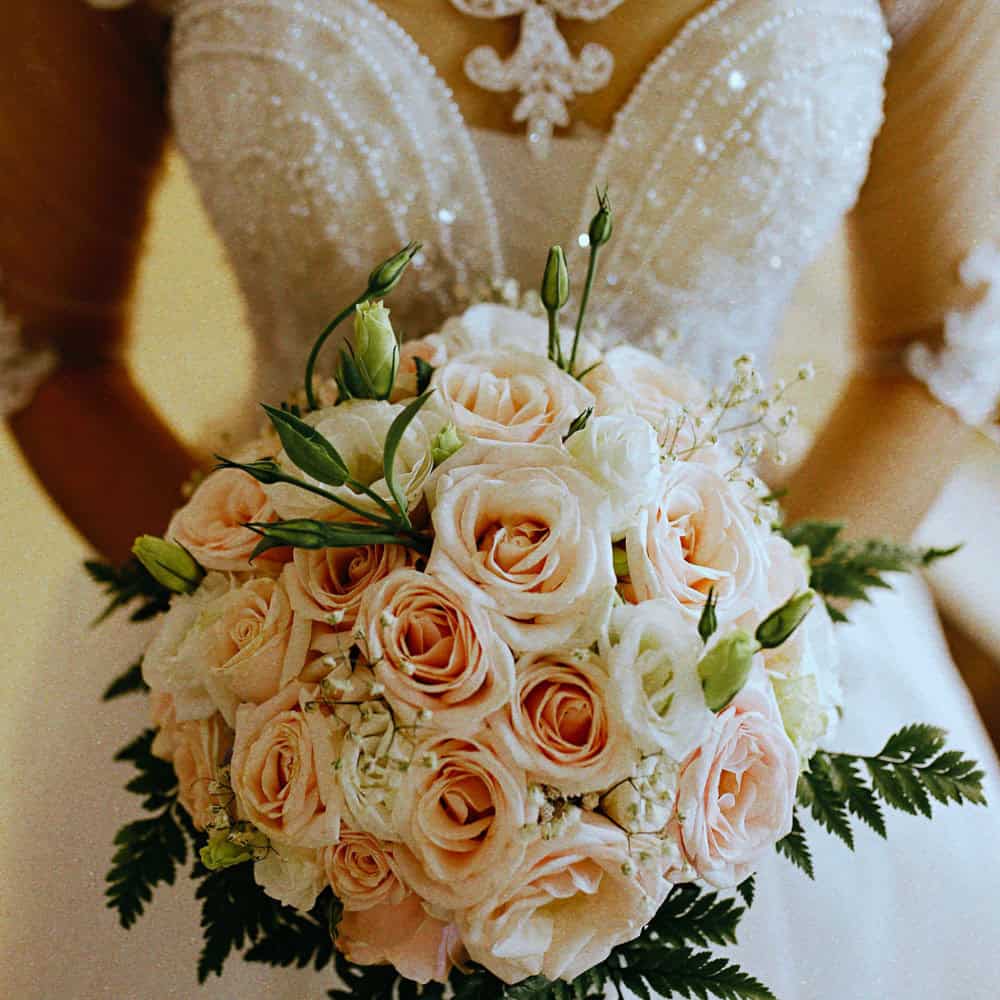A bride in a lace wedding dress holds a bouquet of pale pink and white roses with green foliage.