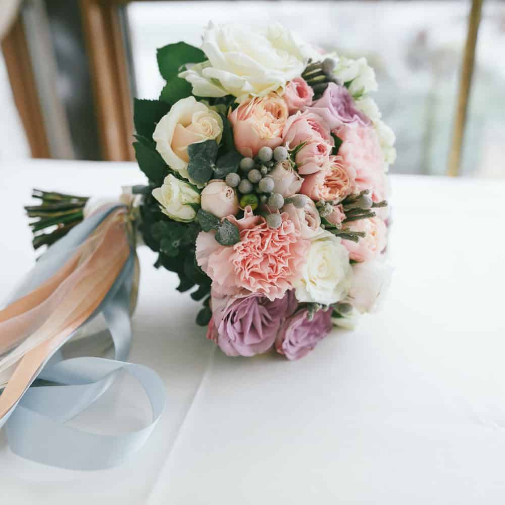 A bouquet of pastel roses, carnations, and greenery with long blue and peach ribbons, resting on a white surface by a window.