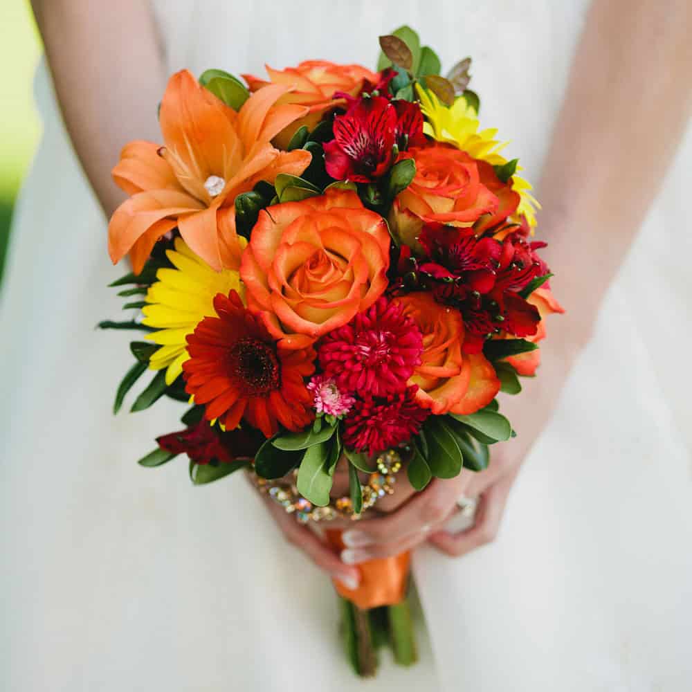 A woman in a white dress holds a bouquet of orange, yellow, and red flowers, including roses, lilies, and daisies.