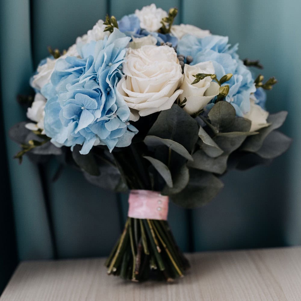 A bouquet of white roses and blue hydrangeas with green leaves, tied together with a pink ribbon, resting on a light-colored surface against a blue curtain.