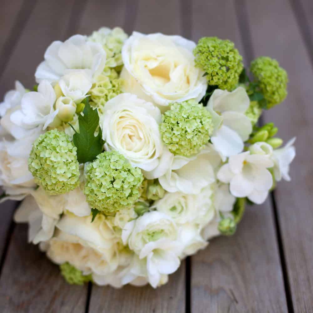 A bouquet of white roses, freesias, and light green hydrangeas is resting on a wooden surface.