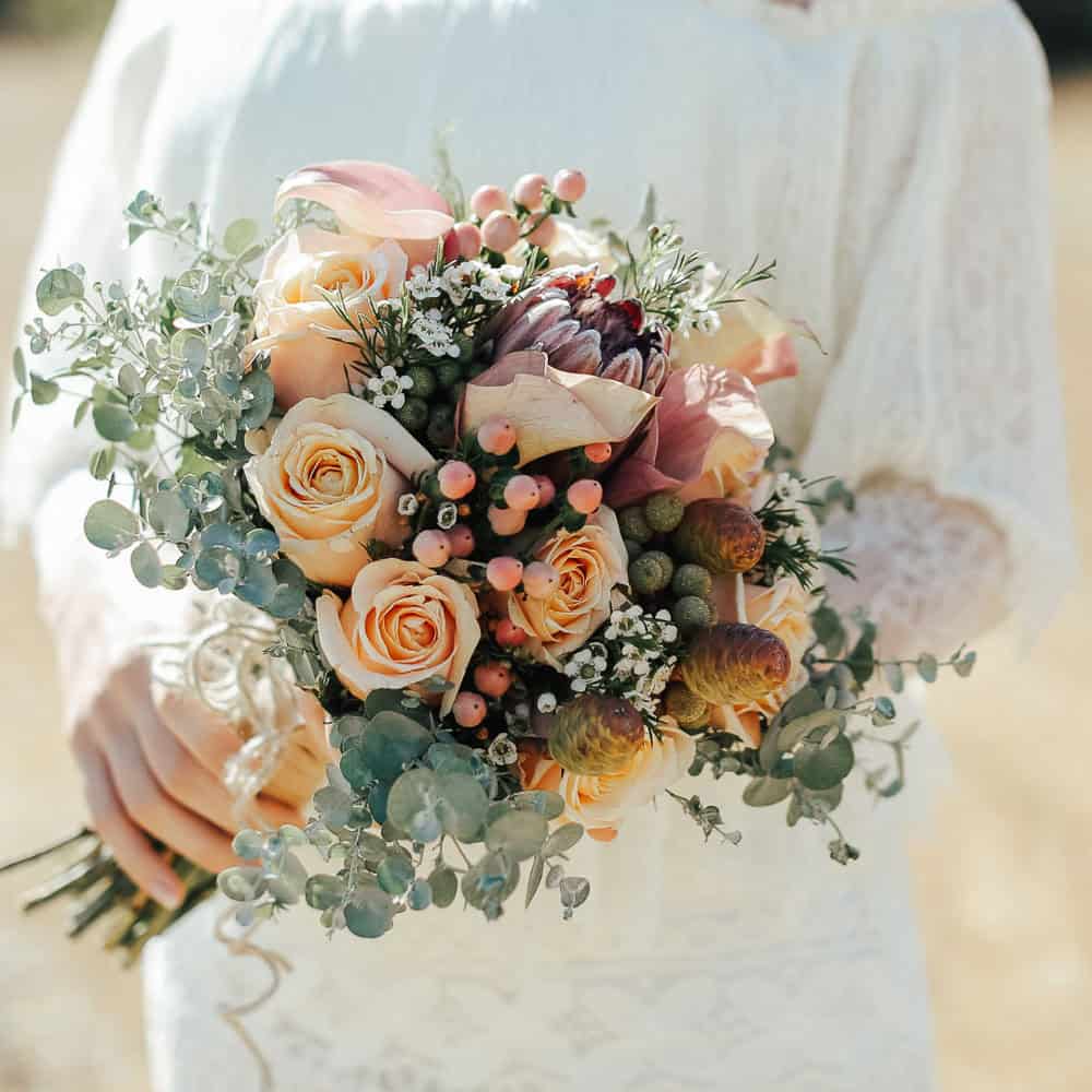 Woman in a white lace dress holding a bouquet with peach roses, eucalyptus leaves, pink berries, and mixed flowers, photographed outdoors.