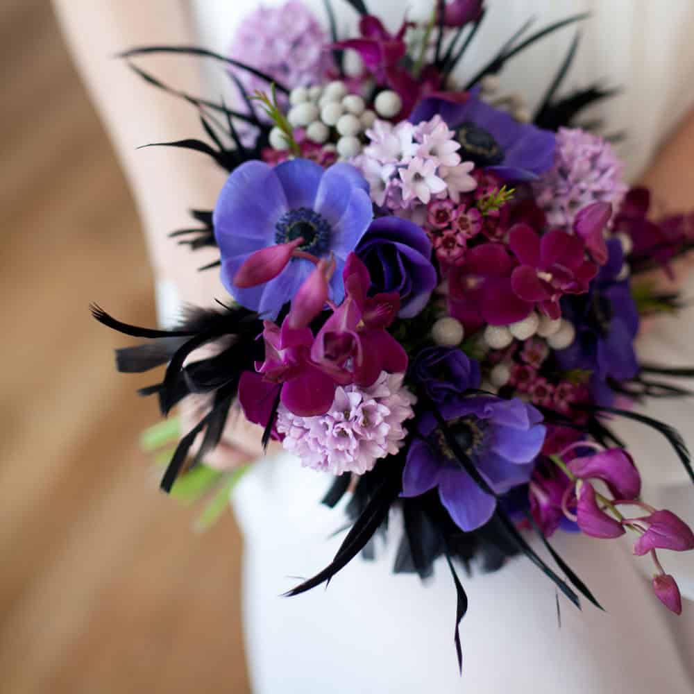 A woman holds a bouquet of purple, violet, and magenta flowers with black accents, set against a blurred background.