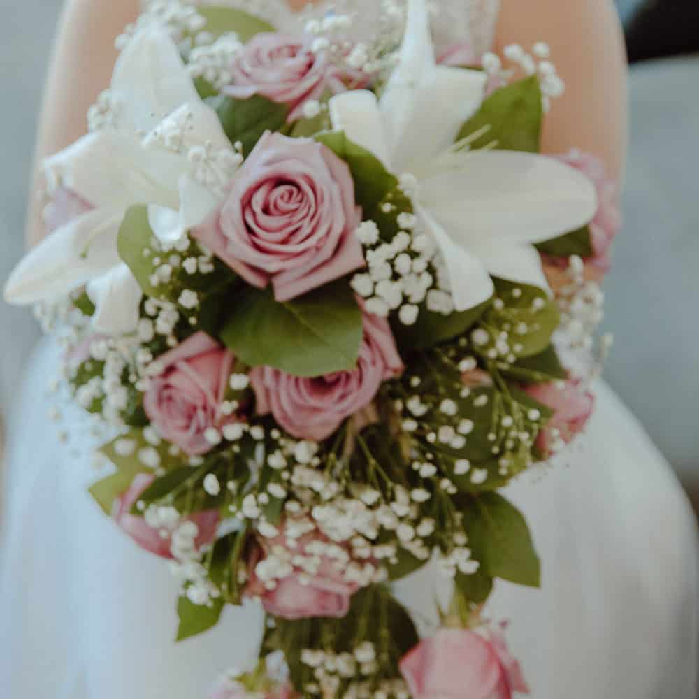 A bridal bouquet featuring pink roses, white lilies, baby’s breath, and green leaves, held by someone in a white dress.