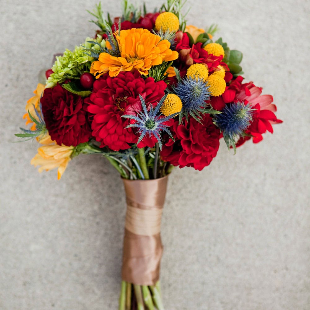A bouquet of red, yellow, and orange flowers with greenery, wrapped with a brown ribbon, displayed against a plain light background.
