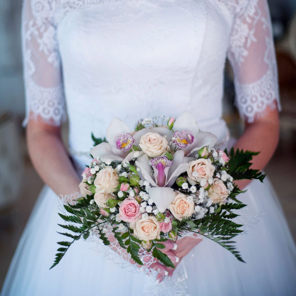 A bride in a white lace wedding dress holds a bouquet of pale pink roses, white orchids, baby's breath, and green ferns.