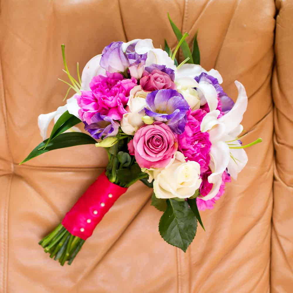 A bouquet of pink, purple, and white flowers with green leaves, wrapped in a red ribbon, resting on a tan leather surface.