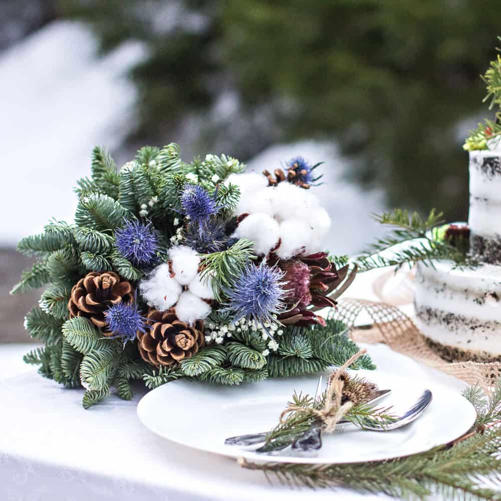 A centerpiece of pine branches, cotton, blue thistles, pinecones, and white flowers sits beside a white plate and cutlery wrapped with evergreen sprigs.