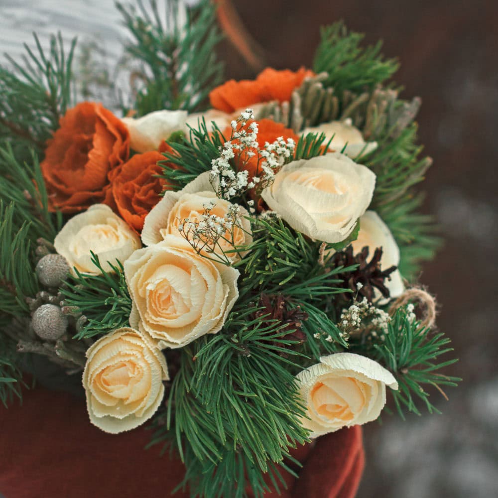 A bouquet featuring cream and orange roses, white baby's breath, green pine branches, and assorted winter foliage.