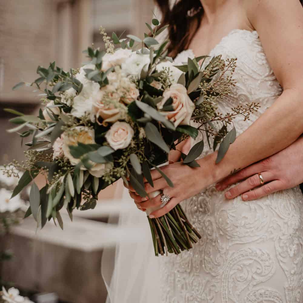 A bride in a white lace wedding dress holds a bouquet of flowers while someone with a gold ring rests their hand on her waist.