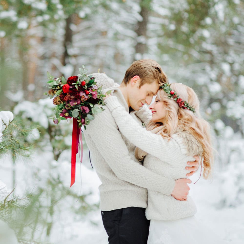 A couple in white sweaters embraces in a snowy forest. The woman holds a bouquet of flowers and wears a floral crown.