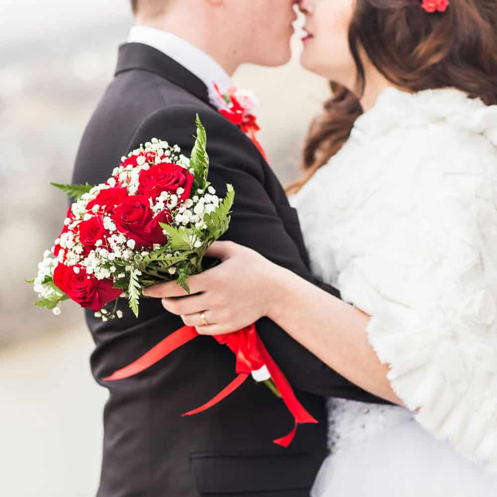 A bride and groom embrace, holding a bouquet of red roses and white baby's breath, dressed in wedding attire.