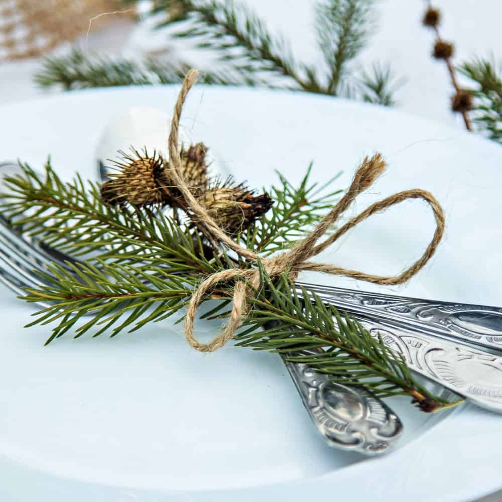 A fork and knife with ornate handles are tied together with twine and decorated with pine sprigs, placed on a white plate.
