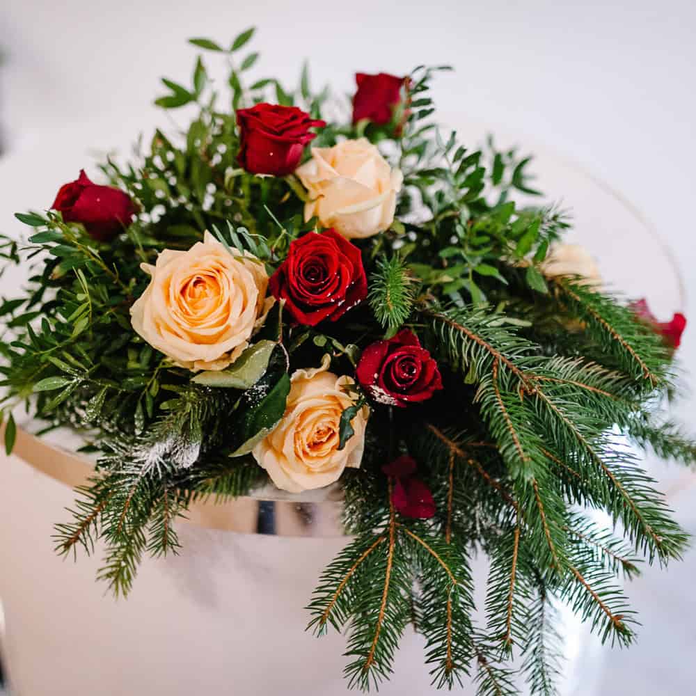 A floral arrangement with red and cream roses, green fir branches, and assorted greenery in a white vase.
