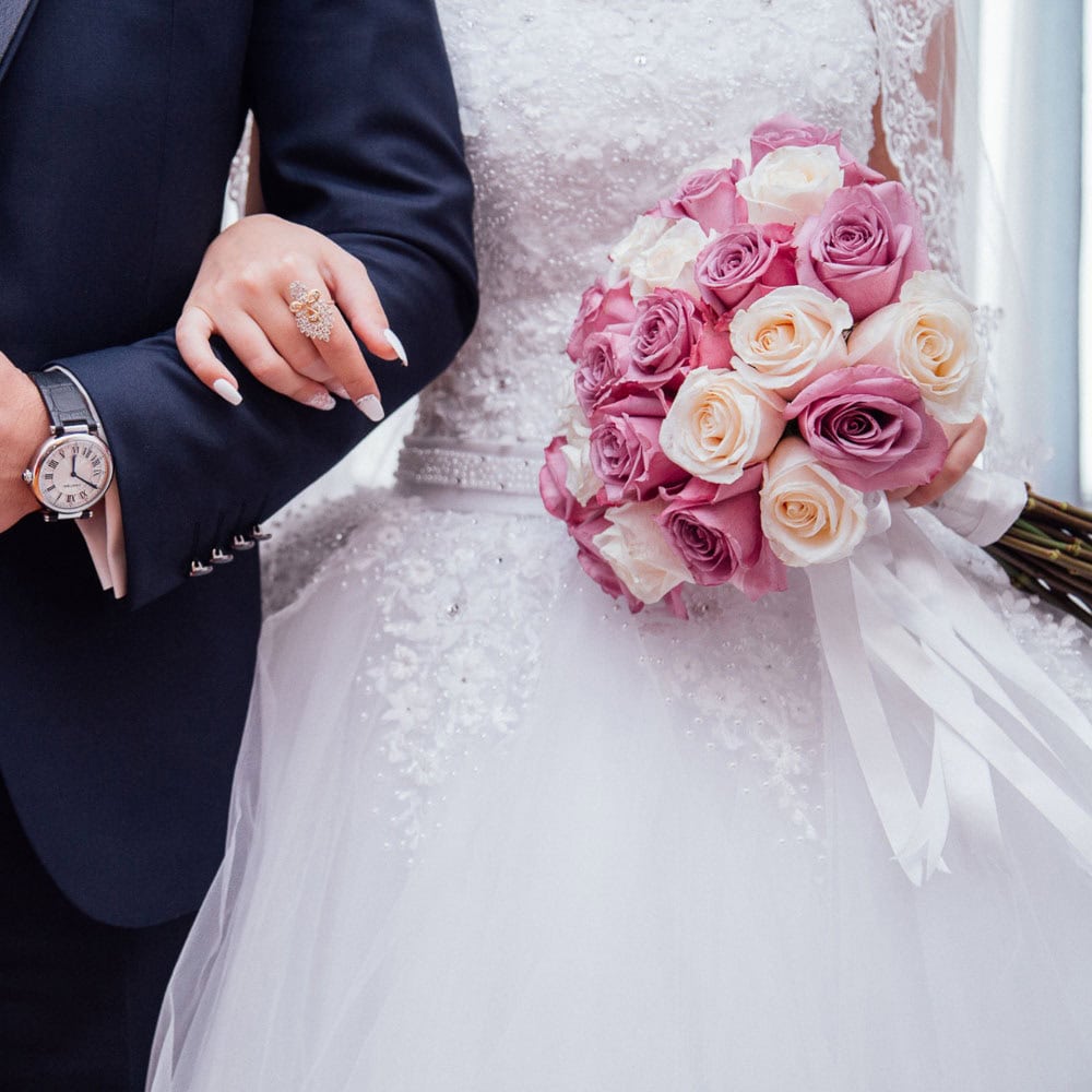 Bride in a white gown holds a bouquet of pink and white roses, standing arm-in-arm with a man in a dark suit wearing a wristwatch.