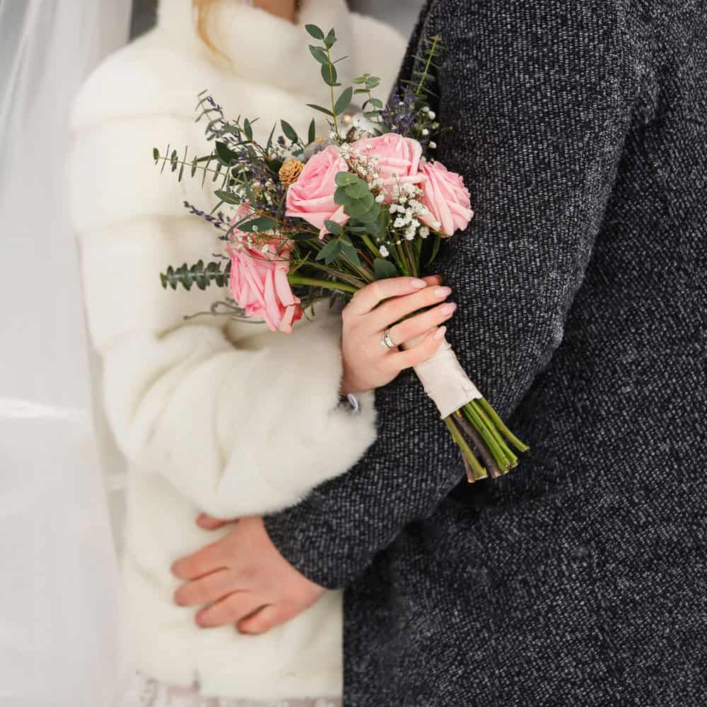 A woman in a white coat holds a bouquet of pink roses and greenery while embracing someone in a dark textured coat.