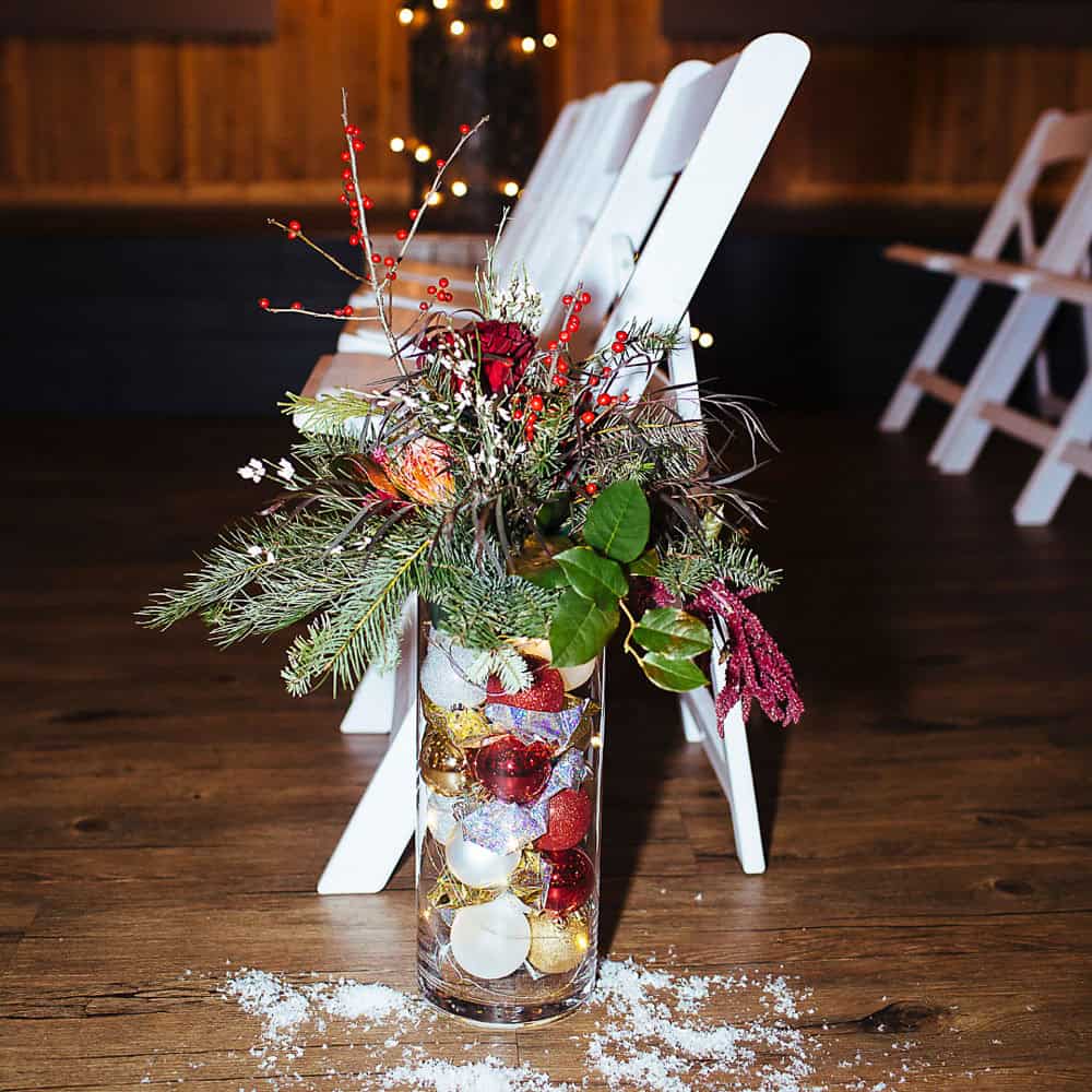 A glass vase filled with Christmas ornaments, pine branches, and red berries is placed on the floor beside white folding chairs on a wooden floor.