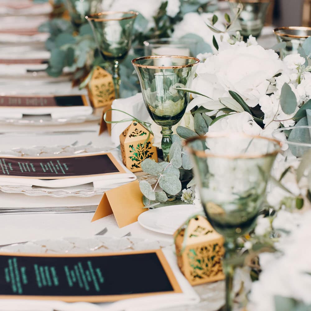 A formal table setting with green glasses, gold-trimmed menus, white floral arrangements, and decorative gold boxes arranged in a row.