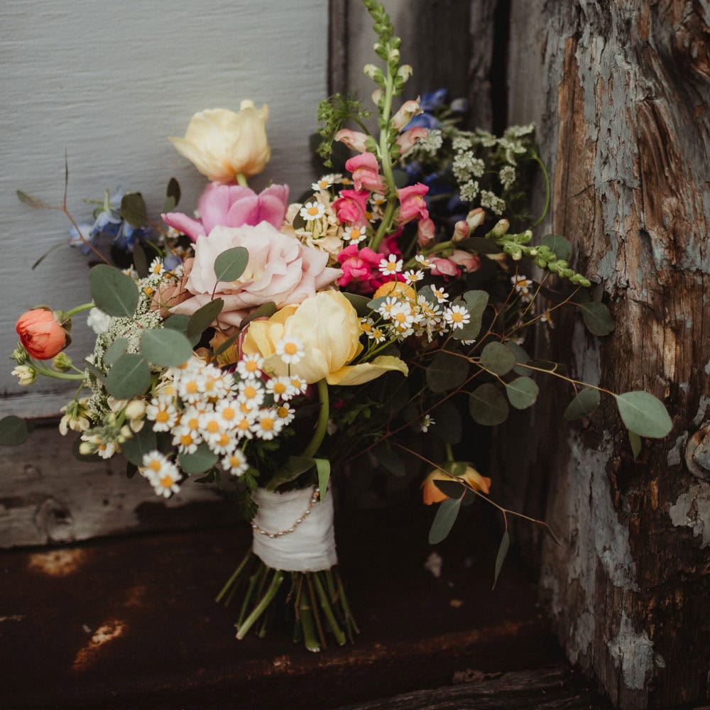 A rustic bouquet of assorted flowers, including daisies, roses, and greenery, is tied with white ribbon and set against a weathered wooden background.
