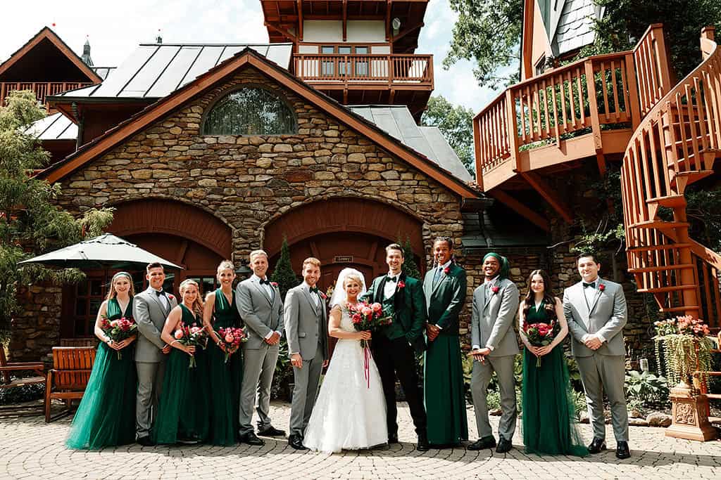 Wedding party poses outdoors before a stone building, with bride and groom centered, surrounded by bridesmaids in green and groomsmen in gray.