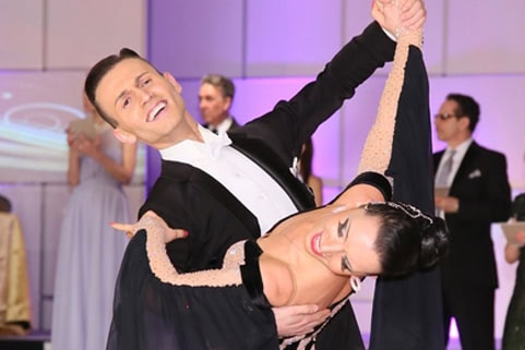 631b8f1508f05e2e4330805b_learn-to-WALTZ-4 A couple in formal attire performs a ballroom dance, with the man holding the woman in a dip pose at a brightly lit indoor event.