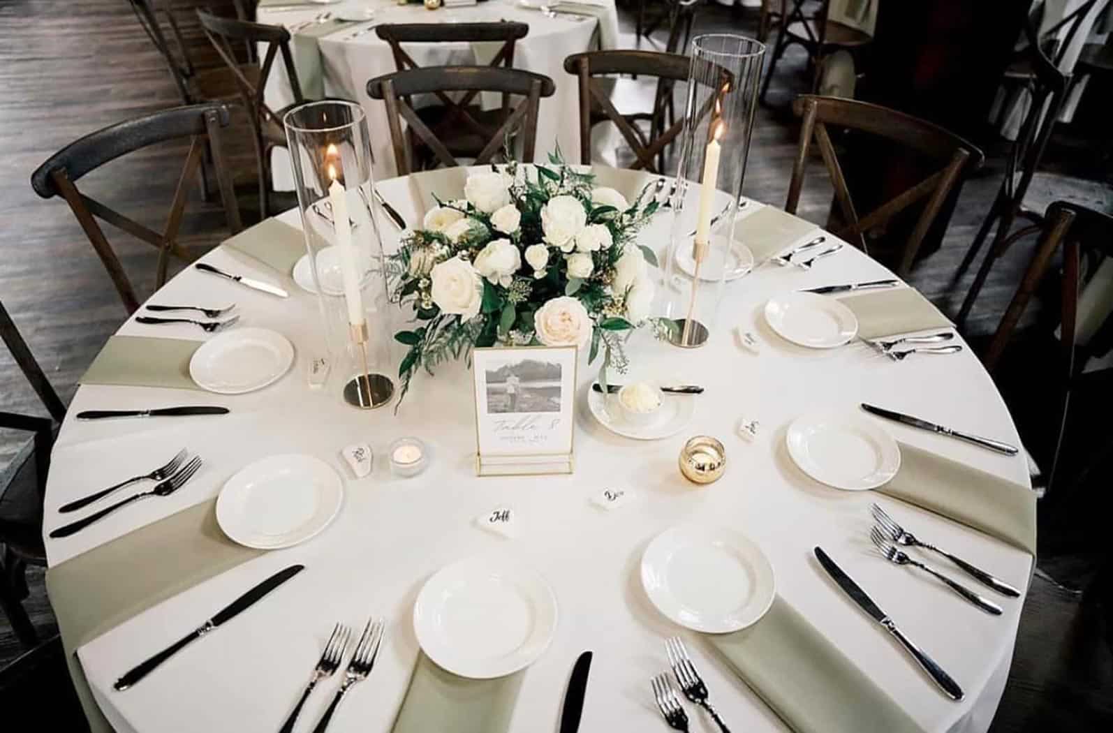 A round table set for eight with white plates, silver cutlery, two tall candles, a white floral centerpiece, and a framed table card on a white tablecloth.