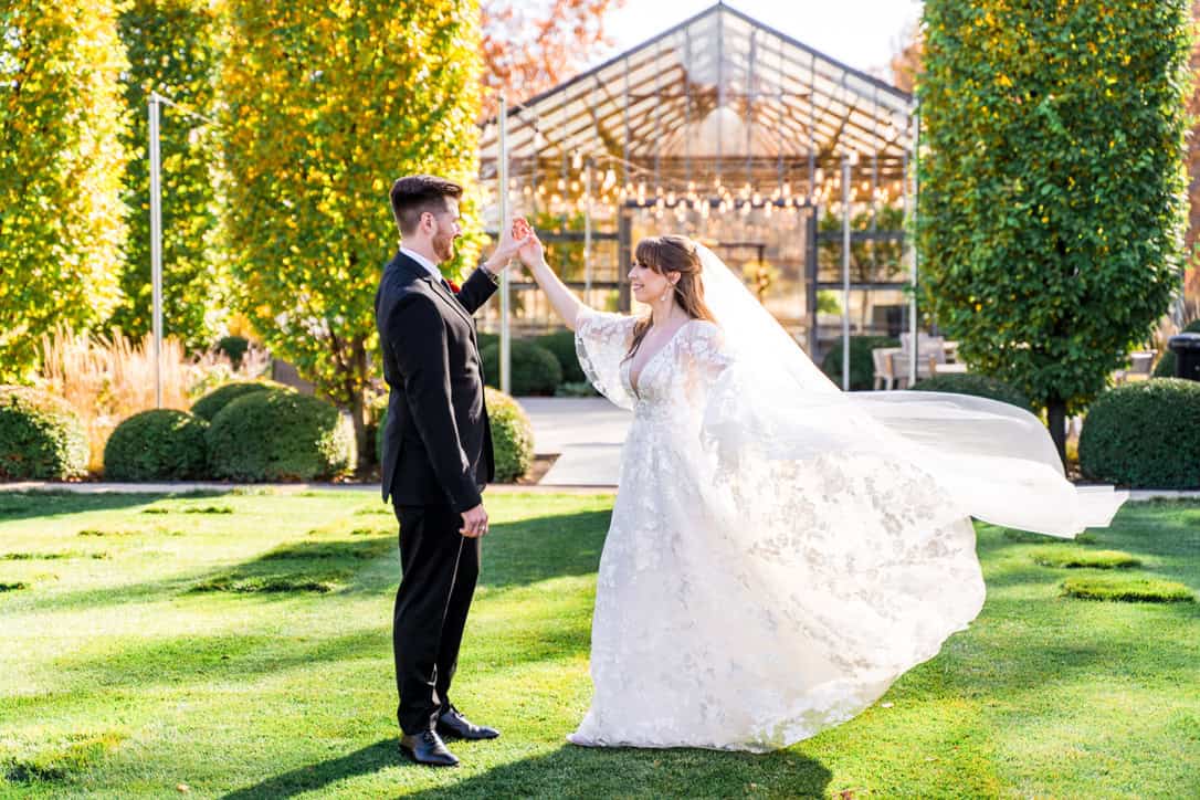 F398A298-3080-4556-9FBD-21F4ABCDD51C_1_105_c A bride and groom stand on grass outside, with the groom twirling the bride; she wears a flowing white dress and veil, and a glass building is in the background.