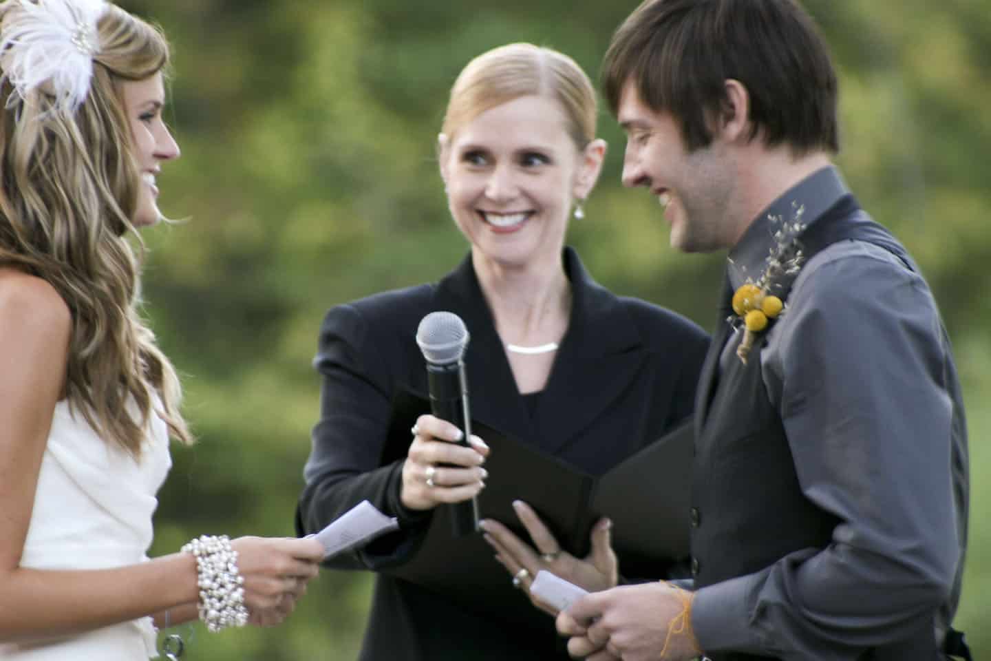 A bride and groom stand facing each other outdoors, holding papers, as an officiant with a microphone smiles at them.