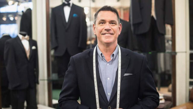 A man in a suit stands in a tailor shop with measuring tape draped around his neck. Mannequins in tuxedos are displayed in the background.