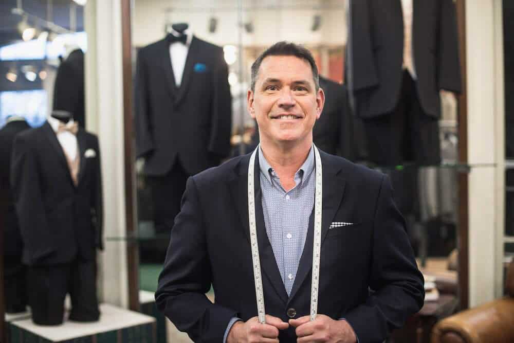 A man in a suit stands in a tailor shop with measuring tape draped around his neck. Mannequins in tuxedos are displayed in the background.