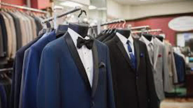 A row of men’s suits, including tuxedos with bow ties and business suits with ties, displayed on mannequins in a clothing store.