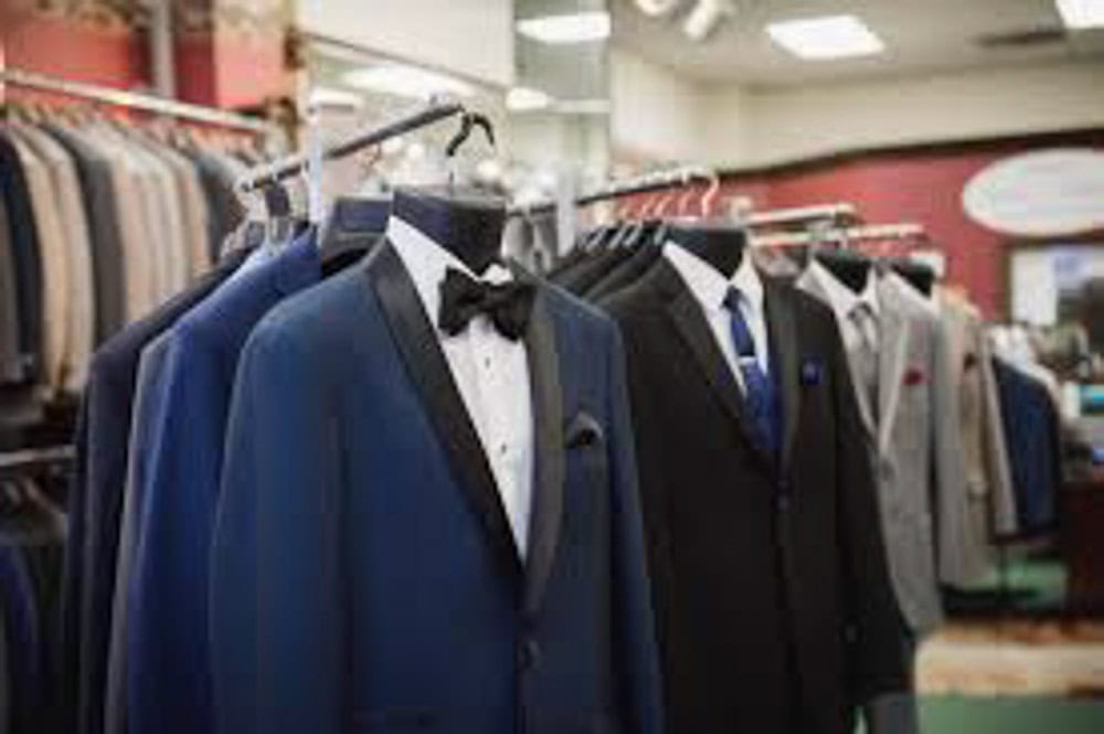 A row of men’s suits, including tuxedos with bow ties and business suits with ties, displayed on mannequins in a clothing store.