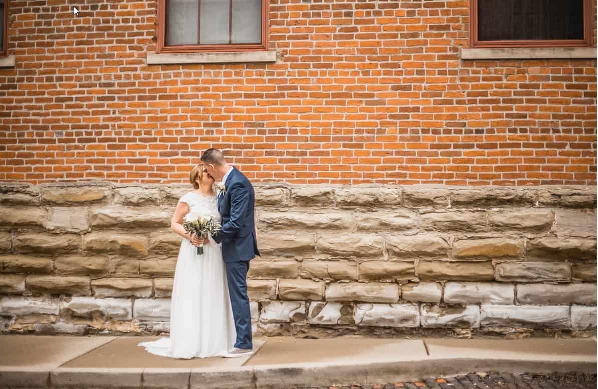 chrome_8tbdGqwYBT A bride and groom stand close together on a sidewalk, kissing in front of a brick and stone wall.