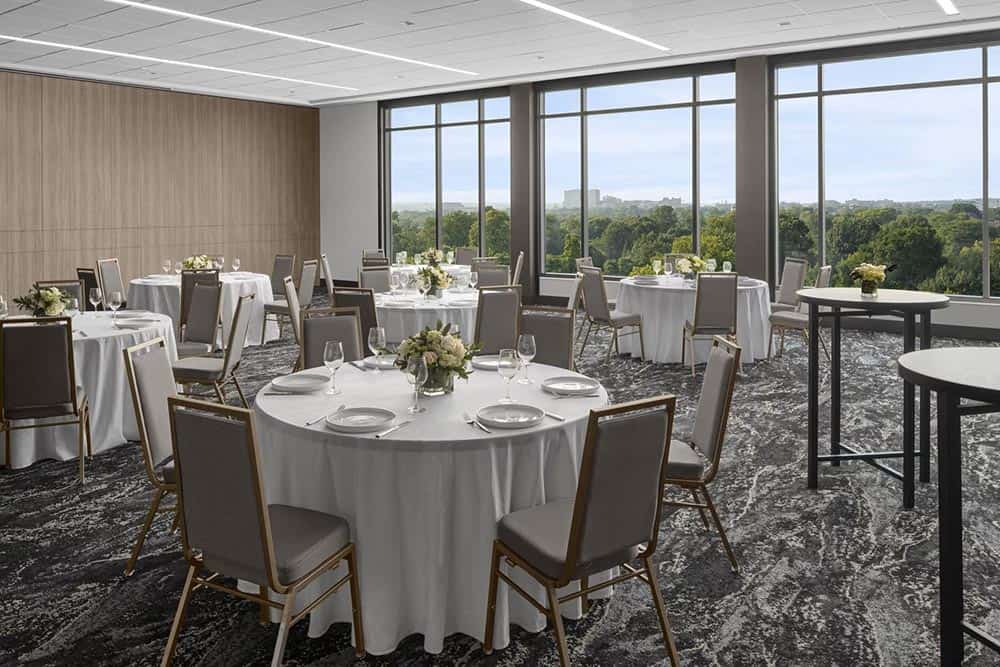 Banquet room with round tables in white tablecloths, set with dishes, glasses, and flower centerpieces, near large windows facing a green landscape.