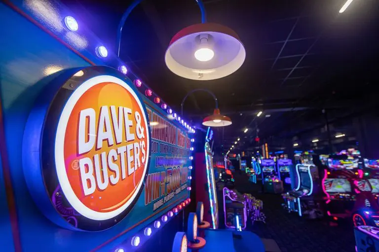 A brightly lit Dave and Buster's sign is displayed on an arcade game, with rows of colorful arcade machines visible in the background.