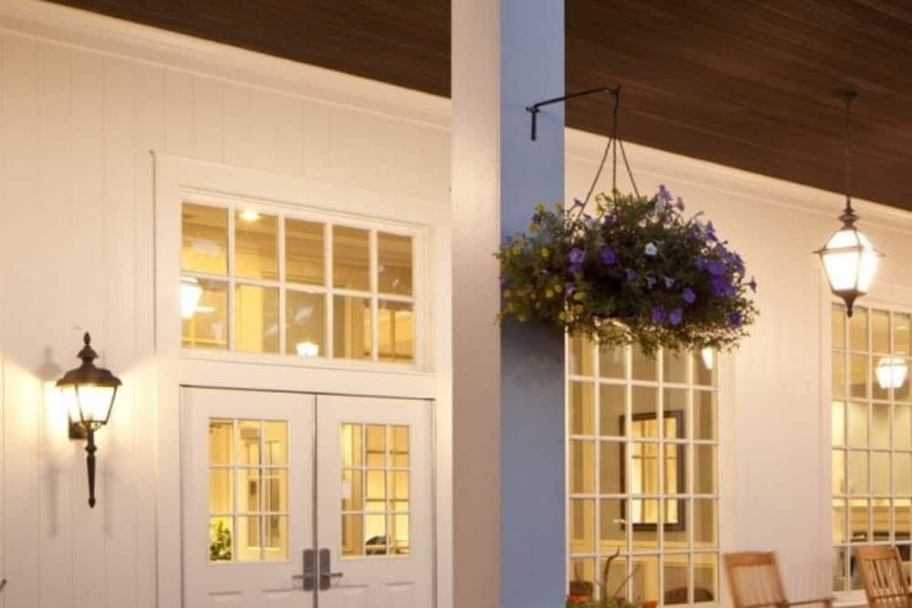White building exterior with double doors, large windows, wall-mounted lanterns, and a hanging basket of purple flowers under a dark wooden porch ceiling.