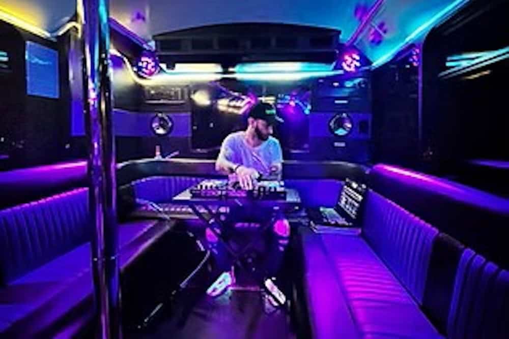A DJ operates mixing equipment inside a party bus with purple and blue LED lighting, surrounded by empty leather seats.