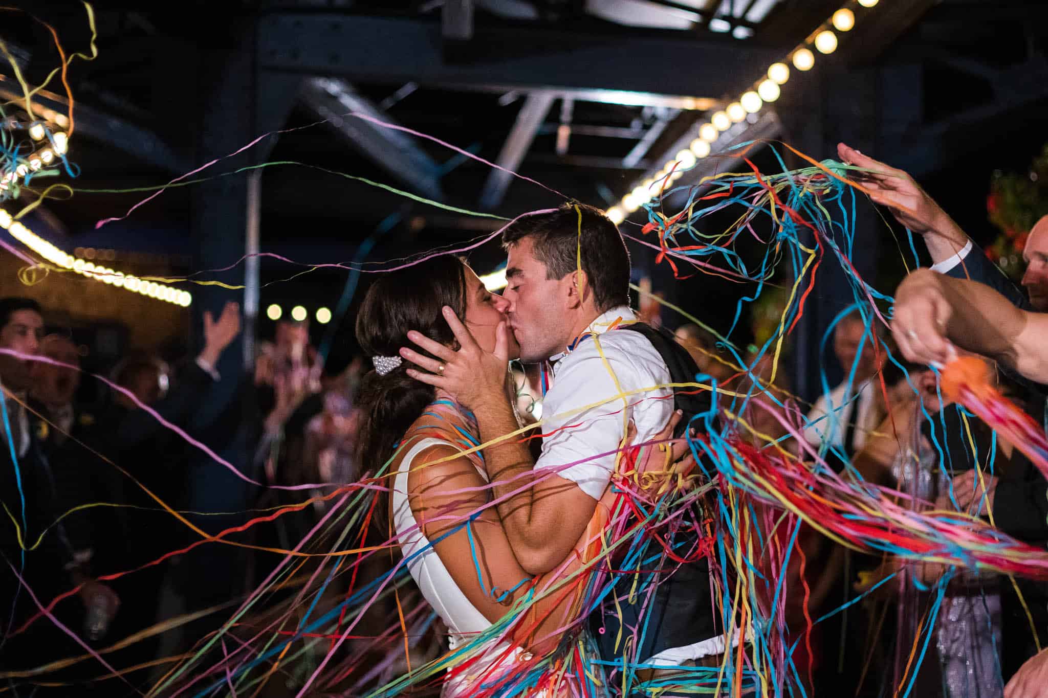 samgracephoto-secondphoto2022-0938 A couple kisses while surrounded by colorful streamers and people celebrating at a festive indoor event.