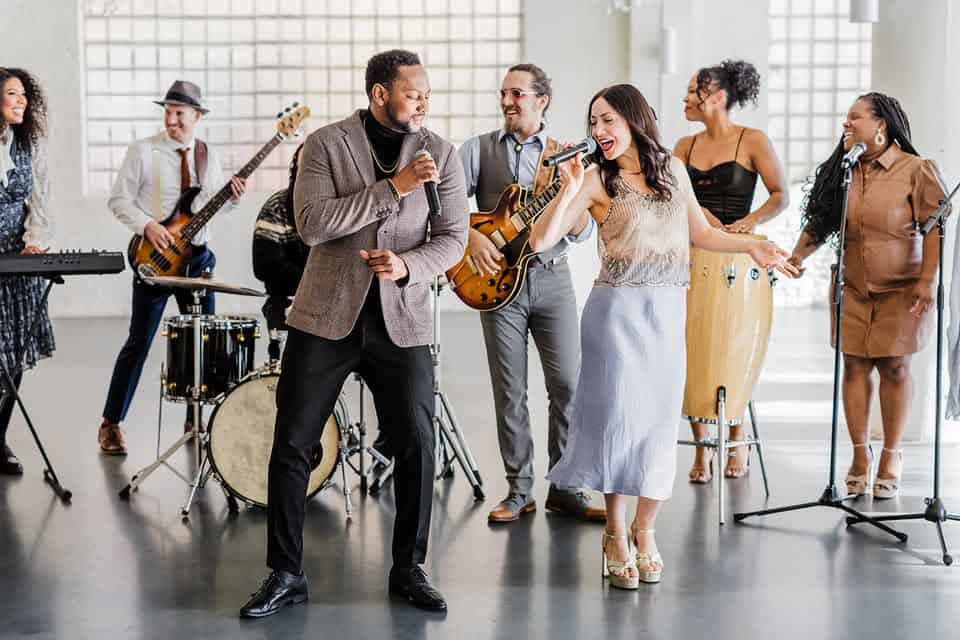 A band of eight musicians perform together indoors, featuring singers, guitar, bass, keyboard, drums, and percussion.