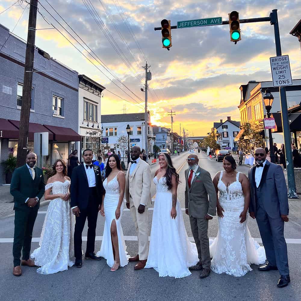 A group of adults dressed in formal wedding attire stands together in the middle of a street at sunset under a Jefferson Ave sign.