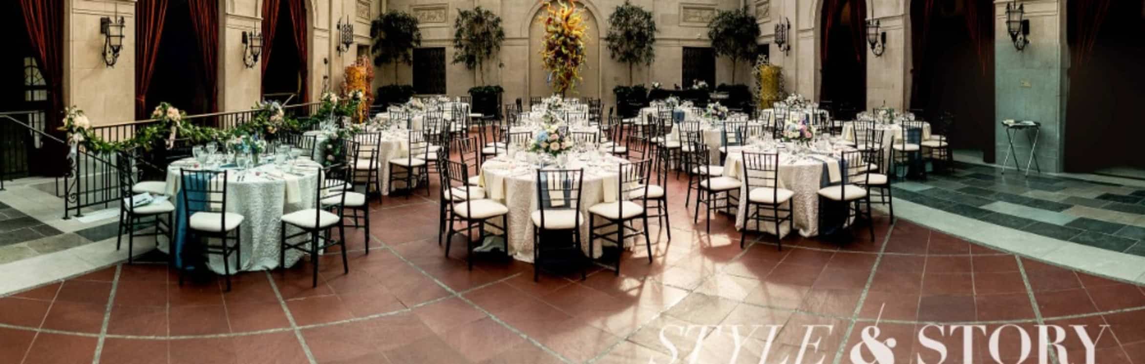 Elegant banquet hall with round tables set for an event, featuring white tablecloths, floral centerpieces, and black chairs on a red and gray tiled floor.