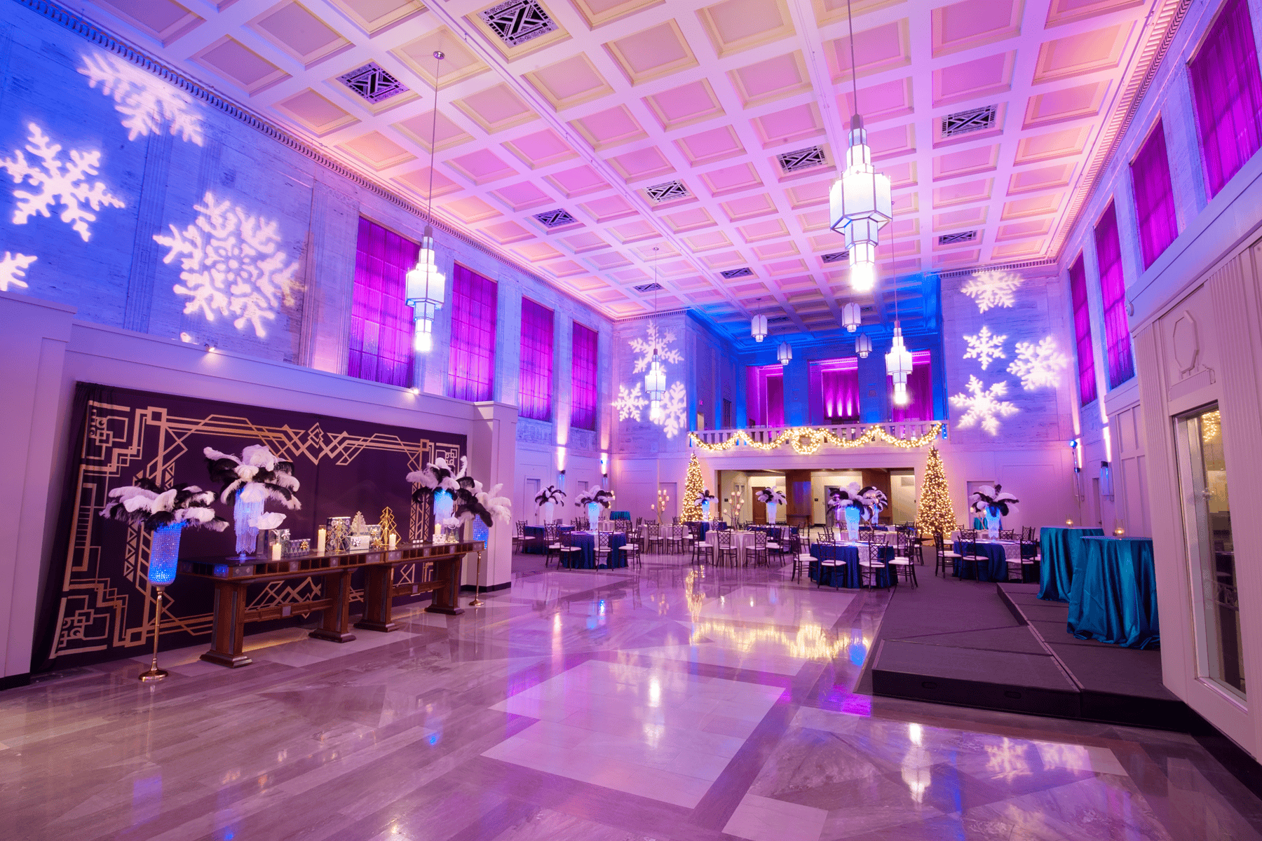 A large banquet hall decorated with snowflake light projections, purple and gold lighting, feather centerpieces, and festive trees, prepared for an event.